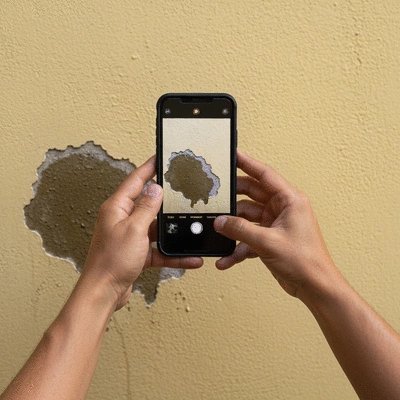 A person's hands holding a smartphone, taking a photo of a damp patch on a wall, documenting housing disrepair, no text, no words, no typography, clean image
